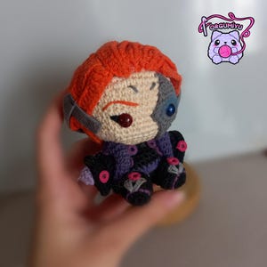 May include: A crocheted plush toy of a character with orange hair, a white face, and a blue eye. The character is wearing a purple and black outfit with pink accents.