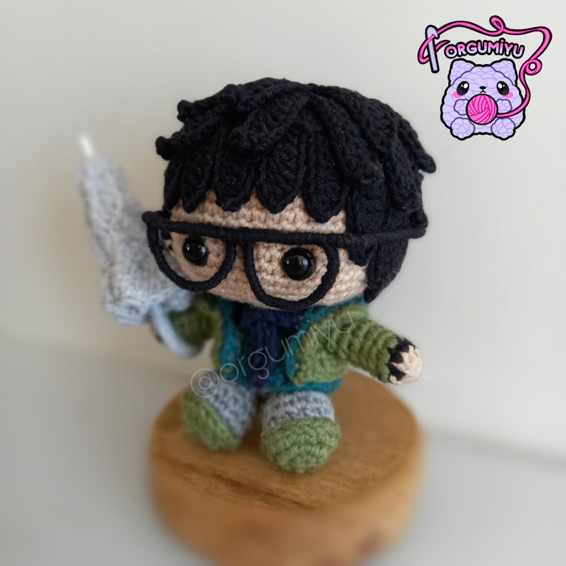 May include: A crocheted amigurumi figure of a person wearing glasses and a green jacket. The figure is holding a crocheted umbrella and is sitting on a wooden base.