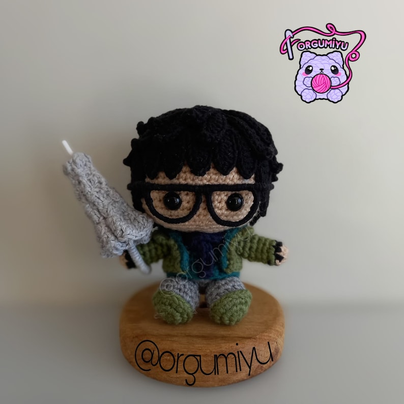 May include: Crocheted amigurumi figure of a person wearing glasses and a green jacket, holding a gray umbrella. The figure is sitting on a wooden base with the text "@orgumiyu" written on it.