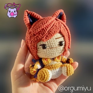 以下が含まれることがあります： 赤い髪、青と黄色の服を着て、猫の耳が付いたキャラクターの編みぐるみ。おもちゃは手のひらに乗っています。