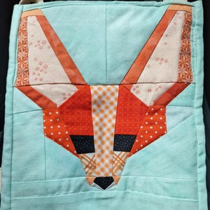 May include: A square, quilted bag featuring a fox face design. The fox is made of orange, white, and black fabric, with a light blue background. The fox's face has a patchwork design with various patterns and textures.