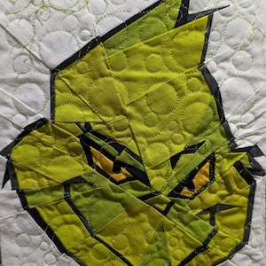 Op de afbeelding: Een gewatteerde textiel met een gestileerde, geometrische afbeelding van een groen gezicht met gele ogen en zwarte accenten. De achtergrond is wit met een herhalend cirkelvormig patroon.