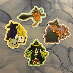 May include: Four Halloween-themed stickers featuring a black cat, a raven, a snake, and a dog. Each sticker has a witch's hat and is illustrated in a cartoon style.