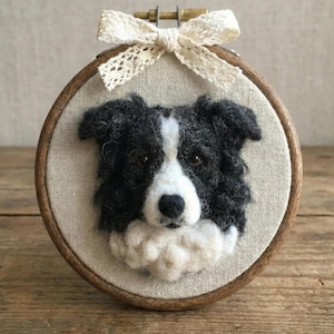 May include: A handcrafted, round embroidery hoop featuring a felted Border Collie dog portrait. The dog has black and white fur, with a cream-colored bow at the top. The hoop is set against a neutral fabric background and a wooden frame.