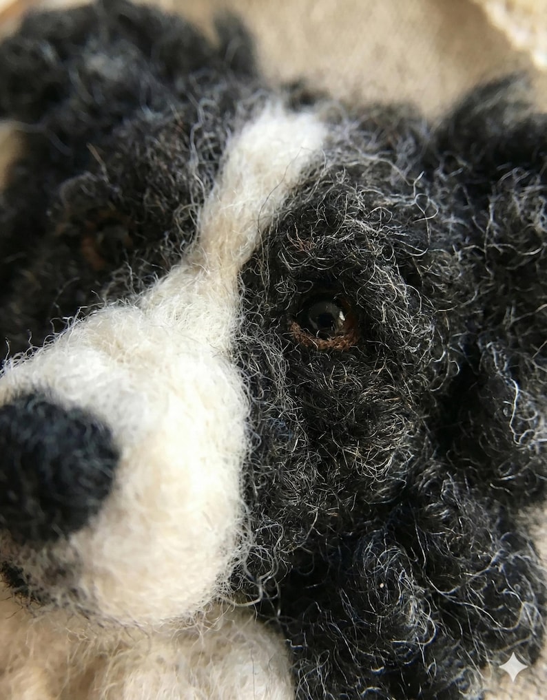 May include: Close-up of a felted dog head with black, white, and gray fur. The dog has a white stripe down its face and a dark brown eye. The fur appears textured and detailed, giving the impression of a handmade craft item.