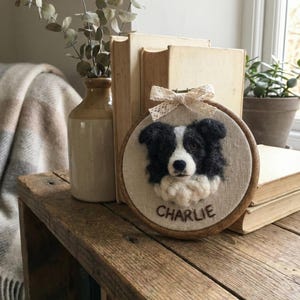May include: A framed textile art piece featuring a black and white dog portrait. The dog's name, "CHARLIE," is embroidered below the image. The artwork is set in a wooden hoop and adorned with a lace bow.