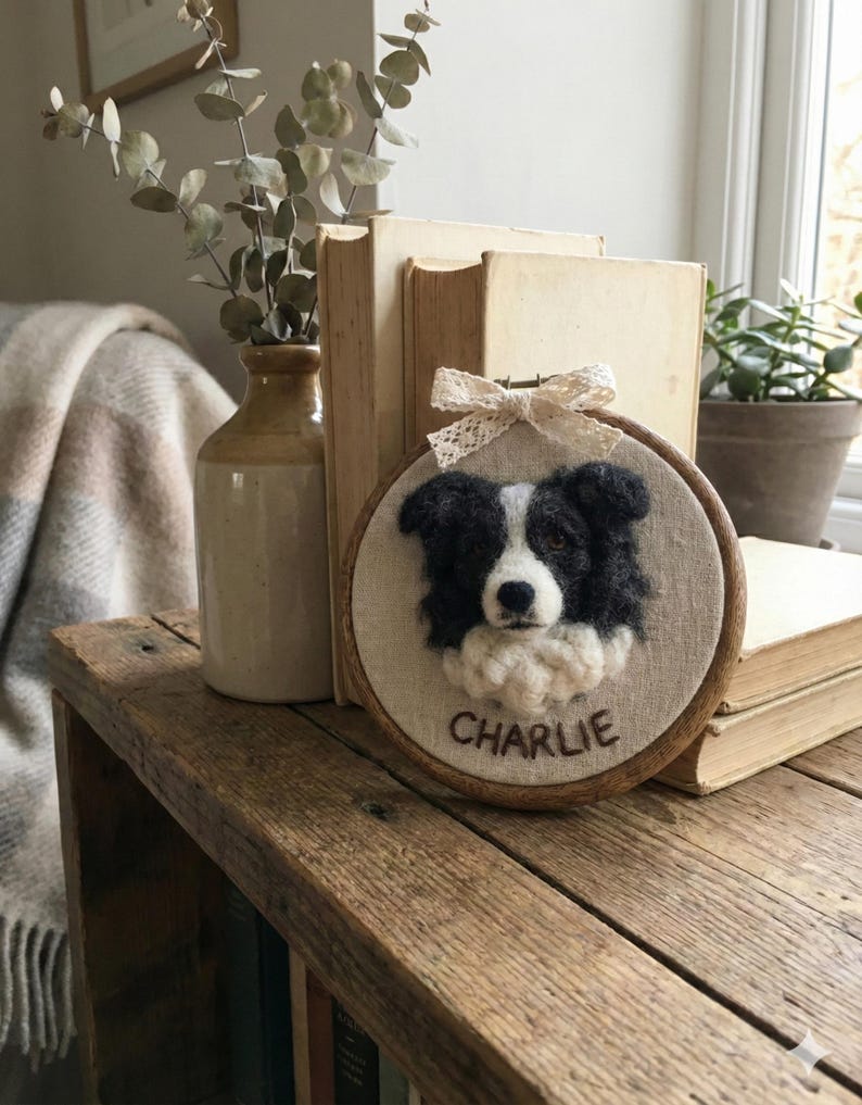 May include: A framed textile art piece featuring a black and white dog portrait. The dog's name, "CHARLIE," is embroidered below the image. The artwork is set in a wooden hoop and adorned with a lace bow.