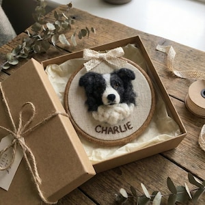 May include: A handmade, framed embroidery of a black and white Border Collie dog named "Charlie." The artwork is presented in a round wooden hoop, adorned with a cream-colored bow, and displayed inside a brown gift box with a twine tie.