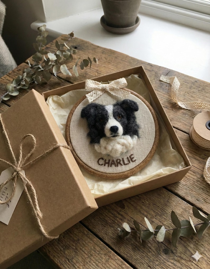 May include: A handmade, framed embroidery of a black and white Border Collie dog named "Charlie." The artwork is presented in a round wooden hoop, adorned with a cream-colored bow, and displayed inside a brown gift box with a twine tie.