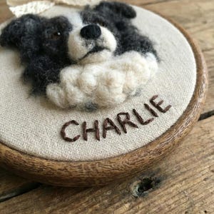May include: A handcrafted, round wall hanging featuring a felted Border Collie dog portrait. The dog is depicted in black, white, and cream colors, with the name "CHARLIE" embroidered below. A cream-colored bow is attached at the top. The artwork is set in a wooden embroidery hoop.