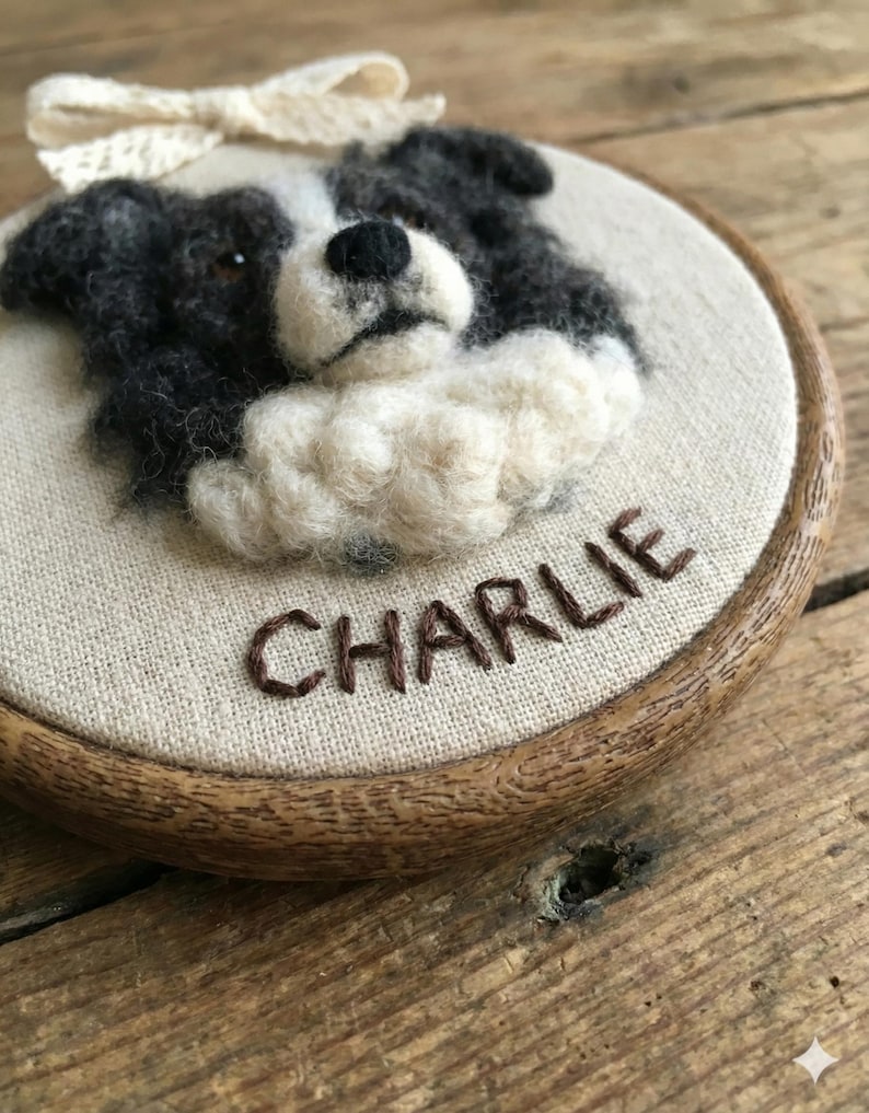 May include: A handcrafted, round wall hanging featuring a felted Border Collie dog portrait. The dog is depicted in black, white, and cream colors, with the name "CHARLIE" embroidered below. A cream-colored bow is attached at the top. The artwork is set in a wooden embroidery hoop.