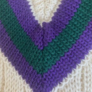 May include: Close-up of a cream-colored knitted garment with a V-neck design. The V-neck features stripes of purple and green. The knitwear has a textured pattern.