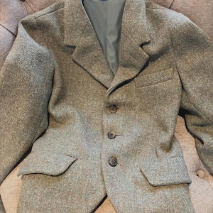 May include: A light brown tweed blazer with a classic three-button closure and two front pockets. The blazer has a notched lapel and a gray lining. The tag inside the blazer reads "MADE IN ENGLAND".