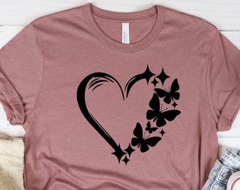 May include: A mauve t-shirt with a black heart design featuring butterflies and stars. The t-shirt is laid flat on a white wooden surface.