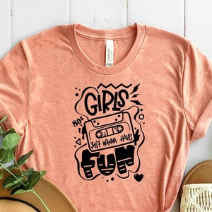 K&ouml;nnte beinhalten: Ein pfirsichfarbenes T-Shirt mit einem schwarzen Grafikdesign, das ein Kassettenband mit dem Text "Girls Just Wanna Have Fun" und der Zahl "80's" zeigt.