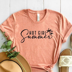 May include: A peach-colored t-shirt with the text "Hot Girl Summer" in black script with palm tree graphics. The shirt is laid out on a white wooden surface with a brown hat and two white sandals.