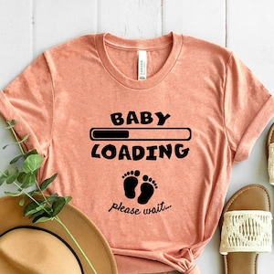 May include: A peach-colored t-shirt with the text "Baby Loading" and a progress bar with two black footprints below. The text "Please wait..." is below the footprints.
