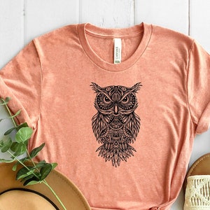 May include: A peach-colored t-shirt with a black and white mandala-style owl graphic. The shirt is made of a soft, comfortable material and has a relaxed fit.