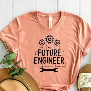 May include: A peach colored t-shirt with the text "FUTURE ENGINEER" in black lettering. The shirt has three black gears above the text and a wrench below the text. The shirt is laid out on a white wooden surface with a brown hat and two white sandals.