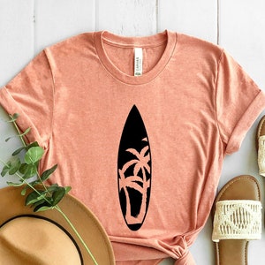 May include: A coral colored t-shirt with a black silhouette of a surfboard with palm trees on it. The shirt is laid out on a white wooden surface with a brown hat and two white sandals.
