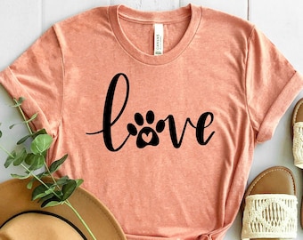 May include: Peach-colored t-shirt with the word "love" in black script, featuring a paw print with a heart. A tan fedora hat and a pair of cream-colored sandals with woven details are also visible.