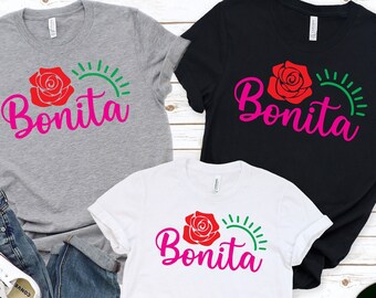 May include: Three t-shirts with a red rose and green sun graphic and the word "Bonita" in pink lettering. The shirts are grey, black, and white.