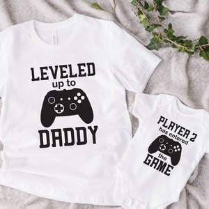 May include: Two white t-shirts with a video game theme. The larger shirt says "LEVELED up to DADDY" with a black video game controller graphic. The smaller shirt says "PLAYER 2 has entered the GAME" with a black video game controller graphic.
