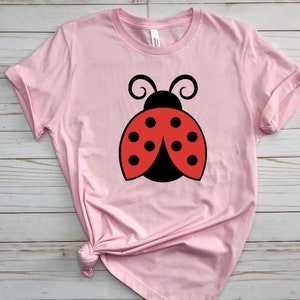 May include: A light pink t-shirt featuring a large cartoon ladybug design. The ladybug has a black body, red wings with black spots, and black antennae. The shirt is made of soft material.