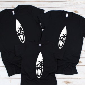 May include: Three black t-shirts with a white silhouette of a surfboard and palm trees. The shirts are arranged in a pyramid shape, with the largest shirt at the top and the smallest shirt at the bottom.