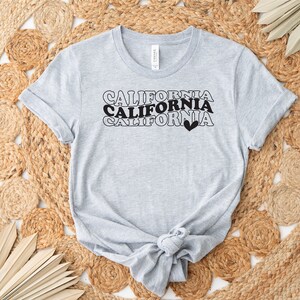 Puede incluir: Una camiseta gris con el texto "CALIFORNIA" repetido tres veces en una fuente retro. El texto es negro y el &uacute;ltimo "CALIFORNIA" tiene un peque&ntilde;o coraz&oacute;n negro a su lado.