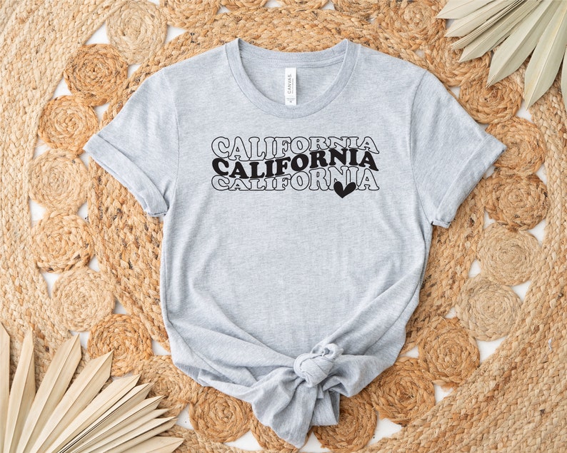 Puede incluir: Una camiseta gris con el texto "CALIFORNIA" repetido tres veces en una fuente retro. El texto es negro y el &uacute;ltimo "CALIFORNIA" tiene un peque&ntilde;o coraz&oacute;n negro a su lado.
