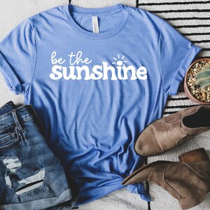 May include: A blue t-shirt with the text "be the sunshine" in white. The shirt is laid out on a white and black striped rug with a pair of denim shorts and brown ankle boots.