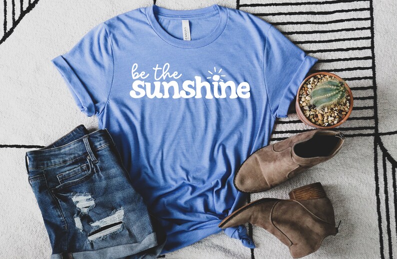 May include: A blue t-shirt with the text "be the sunshine" in white. The shirt is laid out on a white and black striped rug with a pair of denim shorts and brown ankle boots.