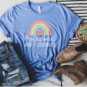 May include: A light blue t-shirt with the text "Professional Anxiety Coordinator" and a rainbow graphic. The shirt is paired with denim shorts and brown ankle boots. A small potted cactus is also visible.