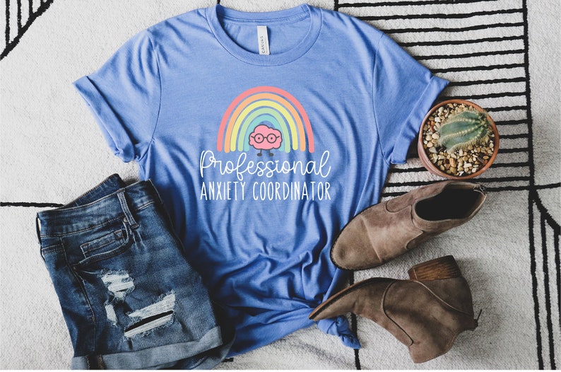 May include: A light blue t-shirt with the text "Professional Anxiety Coordinator" and a rainbow graphic. The shirt is paired with denim shorts and brown ankle boots. A small potted cactus is also visible.