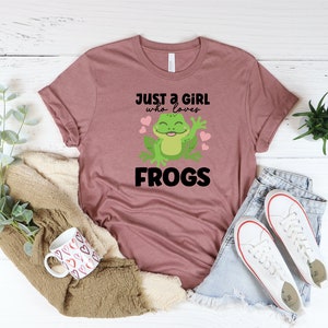 K&ouml;nnte beinhalten: Ein mauvefarbenes T-Shirt mit einem Grafikdruck einer gr&uuml;nen Frosch mit rosa Herzen und dem Text "Just a girl who loves frogs".