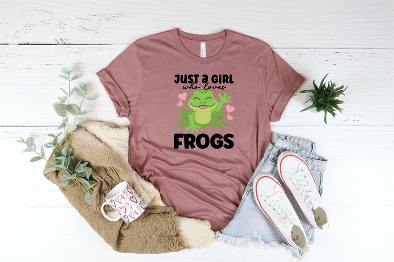 K&ouml;nnte beinhalten: Ein mauvefarbenes T-Shirt mit einem Grafikdruck einer gr&uuml;nen Frosch mit rosa Herzen und dem Text "Just a girl who loves frogs".