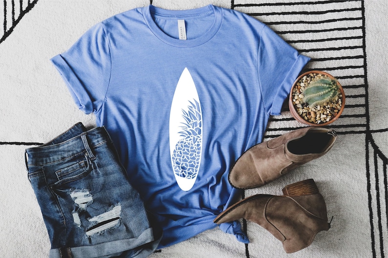 May include: A blue t-shirt with a white pineapple design on a surfboard. The shirt is laid out on a white and black striped rug with a pair of brown ankle boots and a small potted cactus.