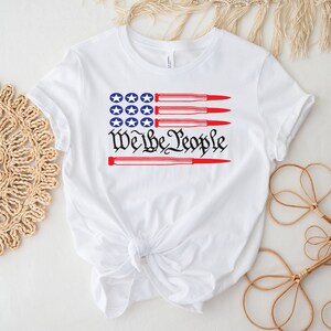 May include: White t-shirt with a red, white, and blue American flag design. The flag is made up of red and white stripes with a blue field of stars. The text "We the People" is printed below the flag, with four red bullets crossing through the text.