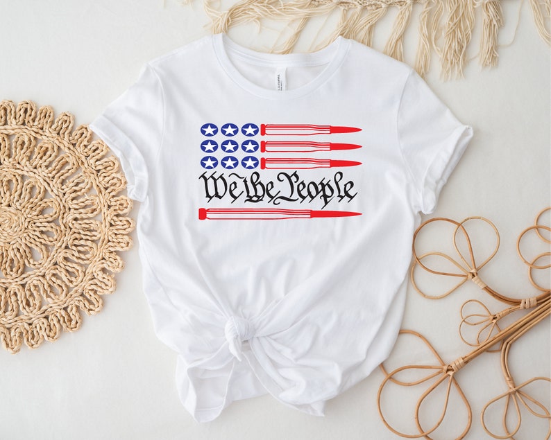 May include: White t-shirt with a red, white, and blue American flag design. The flag is made up of red and white stripes with a blue field of stars. The text "We the People" is printed below the flag, with four red bullets crossing through the text.