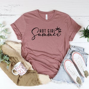 May include: A mauve t-shirt with the text "Hot Girl Summer" in black script with palm tree graphics. The shirt is laid out on a white wooden surface with a mug, a blanket, and a pair of white sneakers.