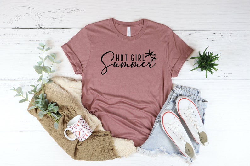 May include: A mauve t-shirt with the text "Hot Girl Summer" in black script with palm tree graphics. The shirt is laid out on a white wooden surface with a mug, a blanket, and a pair of white sneakers.
