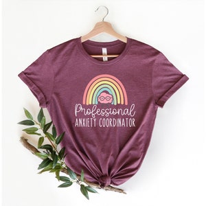 May include: Burgundy t-shirt with rolled sleeves, featuring a rainbow design and the text "Professional Anxiety Coordinator" in white. The rainbow has pastel colors and a cartoon face with glasses. The shirt is hanging on a wooden hanger.