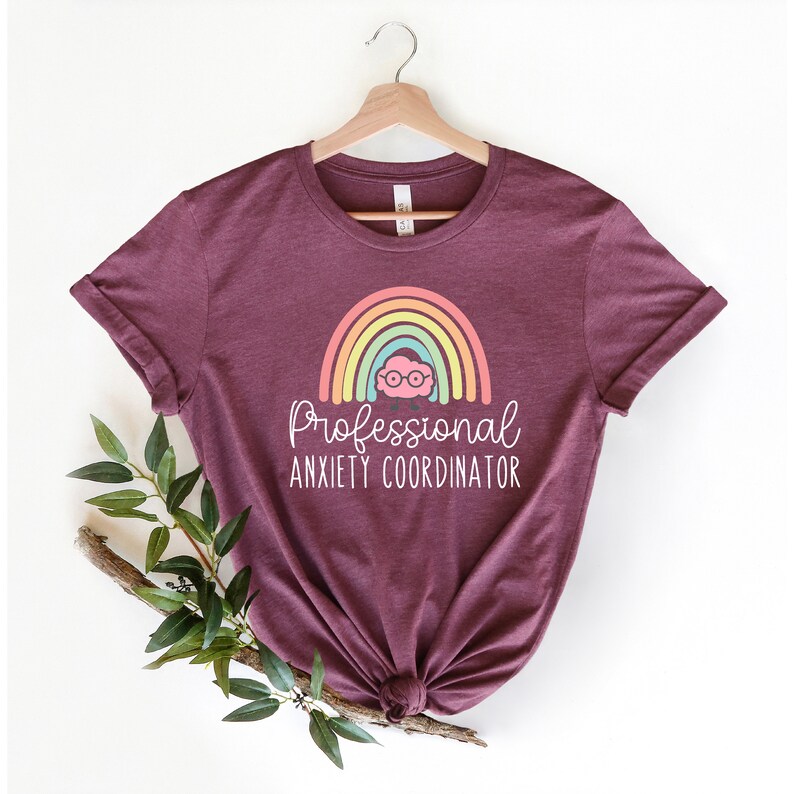 May include: Burgundy t-shirt with rolled sleeves, featuring a rainbow design and the text "Professional Anxiety Coordinator" in white. The rainbow has pastel colors and a cartoon face with glasses. The shirt is hanging on a wooden hanger.