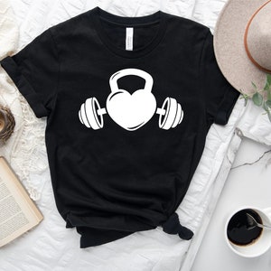 May include: Black t-shirt with a white graphic of a heart with a kettlebell on top and dumbbells on either side.