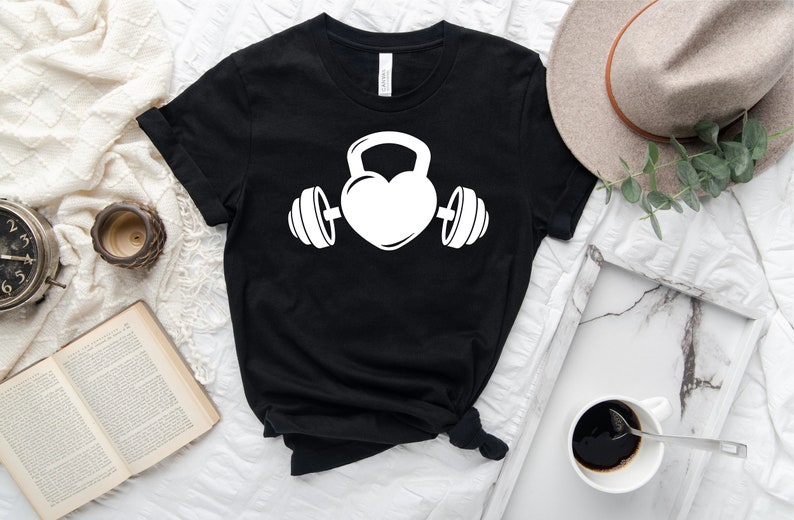 May include: Black t-shirt with a white graphic of a heart with a kettlebell on top and dumbbells on either side.