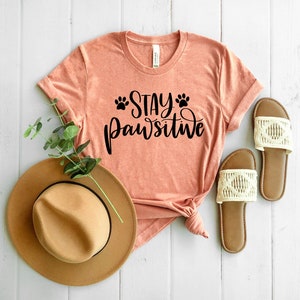 May include: A peach-coloured t-shirt with the text "Stay Pawsitive" in black lettering and paw prints. The shirt is laid out on a white wooden surface with a brown hat and white sandals.