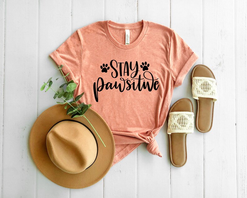 May include: A peach-coloured t-shirt with the text "Stay Pawsitive" in black lettering and paw prints. The shirt is laid out on a white wooden surface with a brown hat and white sandals.