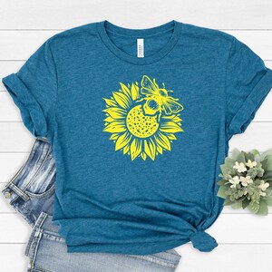 May include: Teal t-shirt featuring a yellow sunflower with a bee graphic. The graphic is centered on the front of the shirt. The shirt is folded and styled with a pair of blue jeans on a white wooden surface.