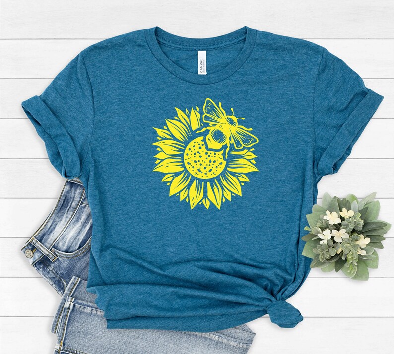 May include: Teal t-shirt featuring a yellow sunflower with a bee graphic. The graphic is centered on the front of the shirt. The shirt is folded and styled with a pair of blue jeans on a white wooden surface.